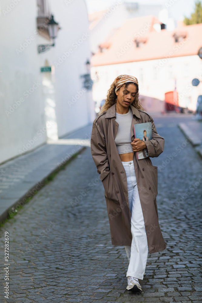 Fototapeta premium full length of african american woman in headscarf and trench coat walking with magazine on street in prague.