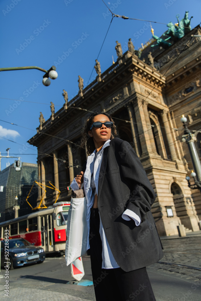 low angle view of young african american woman in stylish blazer and sunglasses posing near national theatre in prague.