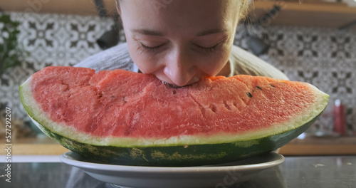 Young girl with braces bites a huge piece of juicy watermelon lying on a plate in the kitchen.