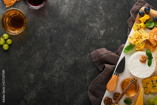 Assortment of cheese, honey, cracker, blueberries, grapes with red and white wine in glasses antipasto server on white marble board on old dark grey background. Flat lay, copy space.