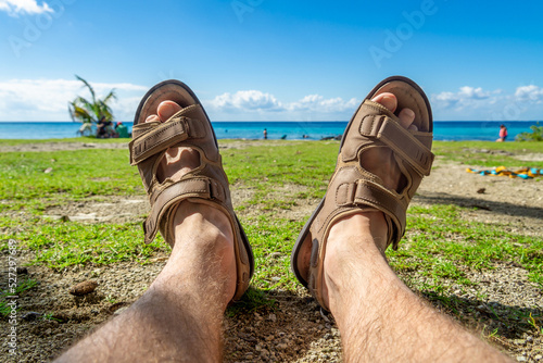Men's feet in sandals against the background of the sea and cloudy sky. A man sunbathes on the shore overgrown with grass. The concept of recreation and tourism.