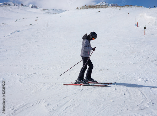 A girl in a ski suit, a helmet and a sun mask stands on skis and with ski poles on a mountain slope against the backdrop of snow-covered mountains.  Winter. Extreme sport and travel content