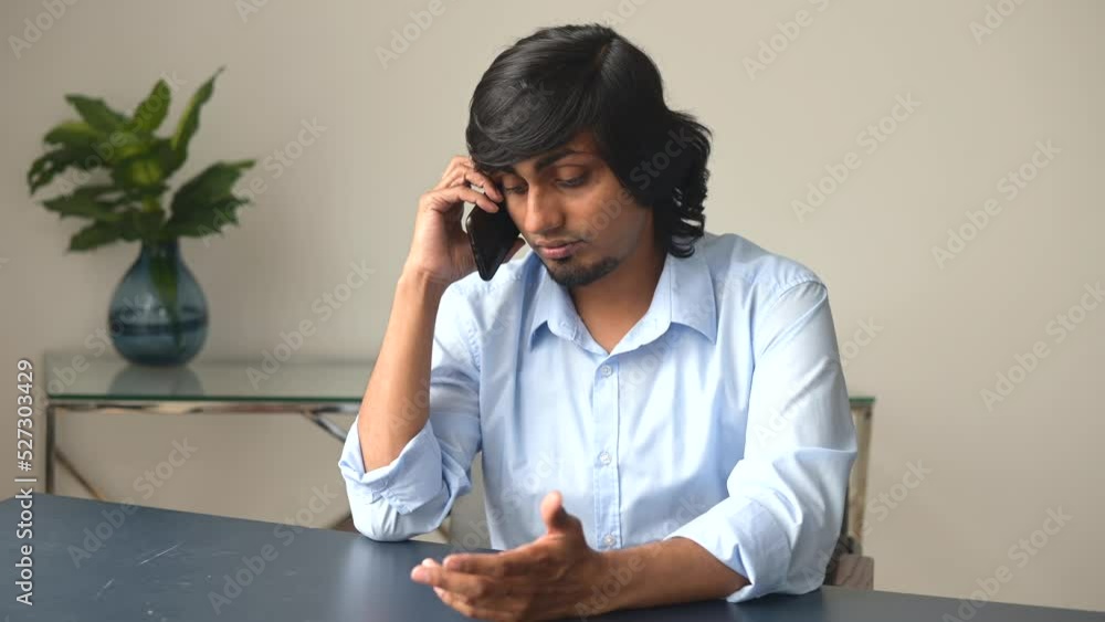 Sad indian businessman in smart casual shirt has unpleasant phone conversation, upset male employee talking on the smartphone sitting at the desk in the office Sad indian businessman in smart casual shirt has unpleasant phone conversation, upset male employee talking on the smartphone sitting at the desk in the office