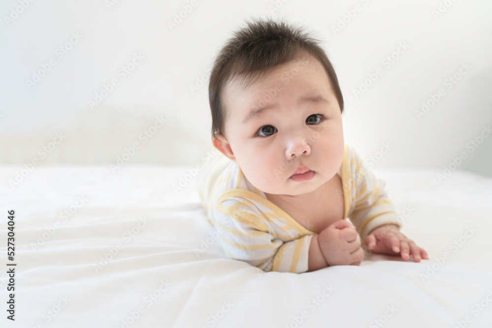 Portrait of cute little asian baby girl wearing bodysuit lying on white bed at home. Cute infant child crawling on bed in the bedroom. Selective focus, free copy space. Childcare Concept. Portrait of cute little asian baby girl wearing bodysuit lying on white bed at home. Cute infant child crawling on bed in the bedroom. Selective focus, free copy space. Childcare Concept.