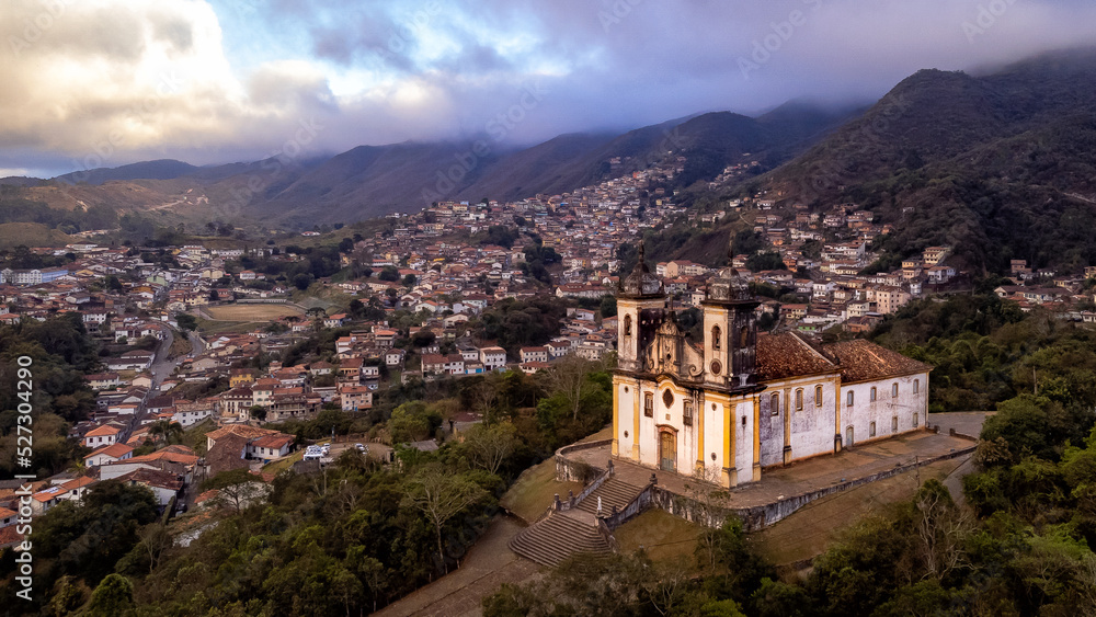 Obraz premium Visão panoramica de igreja em cidade histórica de Ouro Preto Minas Gerais em meio a montanhas e céu nublado construções e casas antigas ao fundo