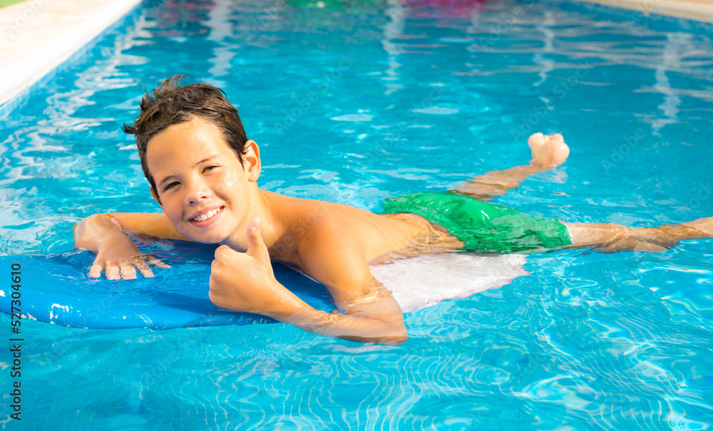 Cute smiling boy paddling on the surf board in pool showing thumb up. Summertime holiday concept.