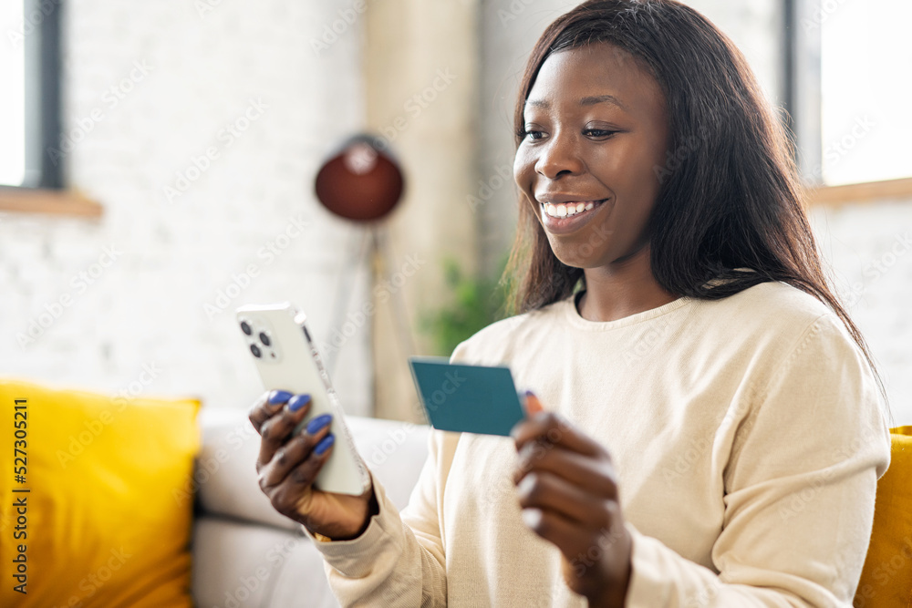 Cheerful young multiracial woman using smartphone shopping app for buying online, holding credit card and inputs banking data, sits on sofa in relaxed atmosphere in living room at home Cheerful young multiracial woman using smartphone shopping app for buying online, holding credit card and inputs banking data, sits on sofa in relaxed atmosphere in living room at home