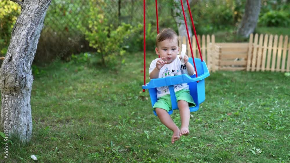 Peaceful Caucasian baby boy swaying in the blue swing in the garden. Trees and wooden fence at backdrop. Peaceful Caucasian baby boy swaying in the blue swing in the garden. Trees and wooden fence at backdrop.