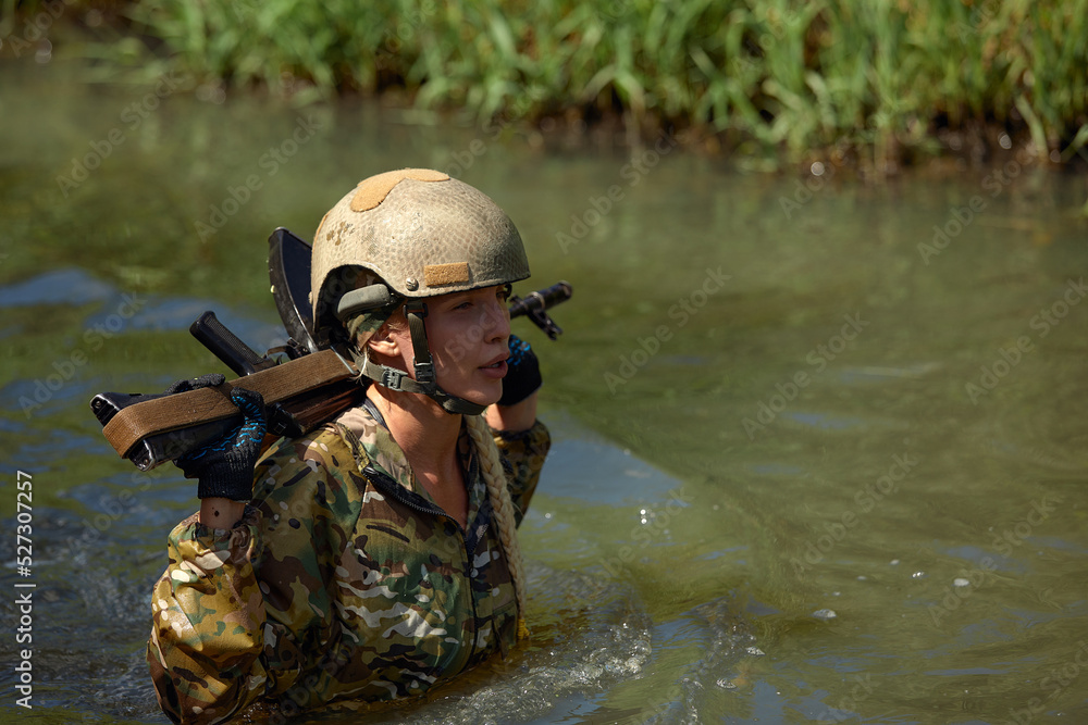 Portrait of fit military female holding weapon rifle in hands in river, posing alone. Confident strong athlete lady looking at side seriously. Attractive good-looking female in military gear