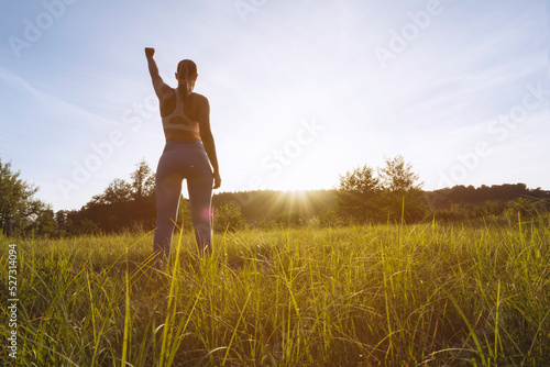 Fototapeta Naklejka Na Ścianę i Meble -  a girl in a fitness suit stands with her back to the camera at sunset with her hand raised up after a workout in the fresh air, motivational girl silhouette there is a place for an inscription