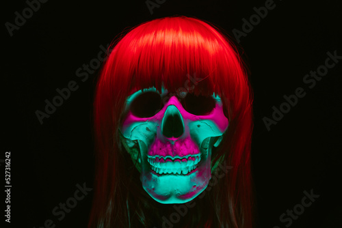 human skull of a woman in a wig with red hair with colored pink green light on a black background