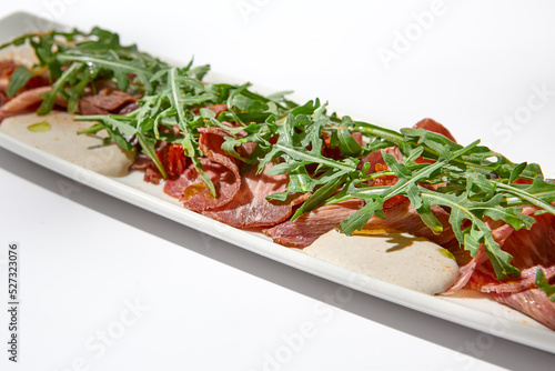 Sliced veal meat on white table with harsh shadows. Sliced veal meat with tuna sauce and arugula. Vitello tonnato on ceramic plate in summer day. Aesthetic food menu photo