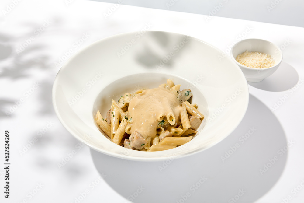 Italian pasta penne with chicken and mushrooms in creamy sauce. Mushroom and chicken pasta on white plate with shadows of sunlight. Summer italian lunch Penne with mushroom sauce on light background