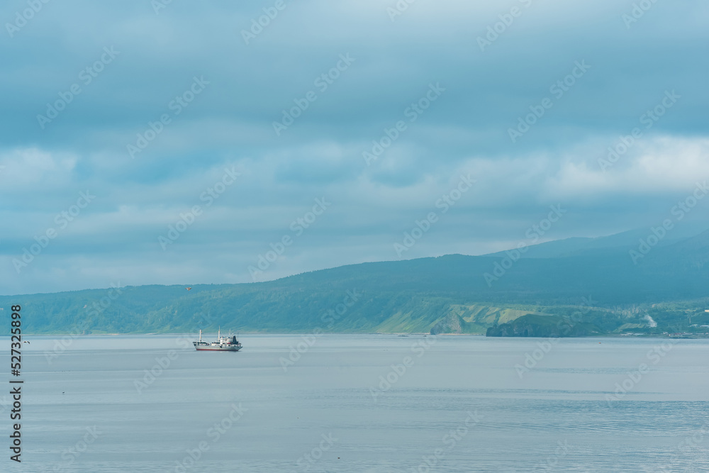 waterscape of the sea bay with a fishing ship and a mount in the clouds in the background, a view of the Mendeleev volcano from the side of the town of Yuzhno-Kurilsk