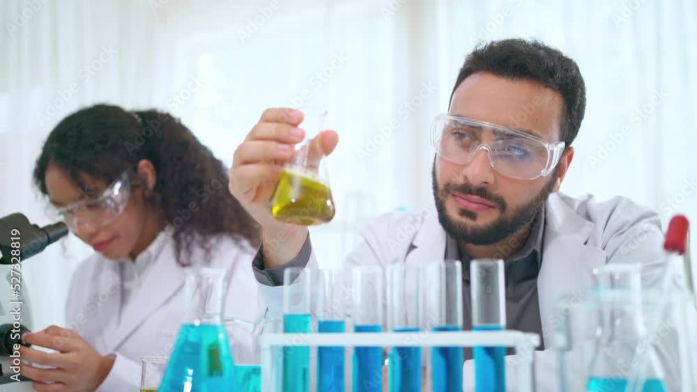 Scientist in lab coat and safety glasses holding flask with yellow solution at modern laboratory. Look at tube and analysis. Research development, bio chemistry, medical and pharmaceutical concept Scientist in lab coat and safety glasses holding flask with yellow solution at modern laboratory. Look at tube and analysis. Research development, bio chemistry, medical and pharmaceutical concept
