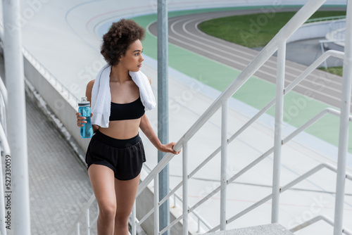Young african american sportswoman with sports bottle walking near railing on stadium.