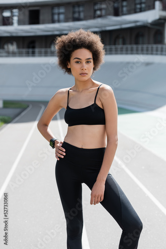 slim african american woman in black sportswear standing with hand on hip and looking at camera.