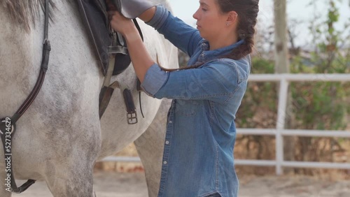 Side view of a young woman putting the saddle on her white horse in a stable