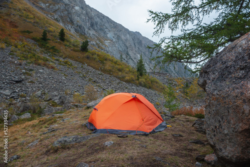 Dramatic mountain landscape with alone orange tent near big rock on hill with autumn flora against large mountain range in overcast. Lonely tent close-up among fading autumn colors in high mountains.