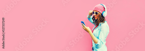 Summer colorful portrait of stylish modern young woman listening to music in headphones with smartphone on pink background