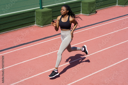 Confident african female athlete running on racetrack. Sportive black sport woman training jog on stadium prepare for competition event, marathon or triathlon. Mix race girl runner practicing outdoor © DimaBerlin