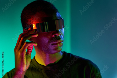 Serious young ethnic man touching VR glasses in neon studio