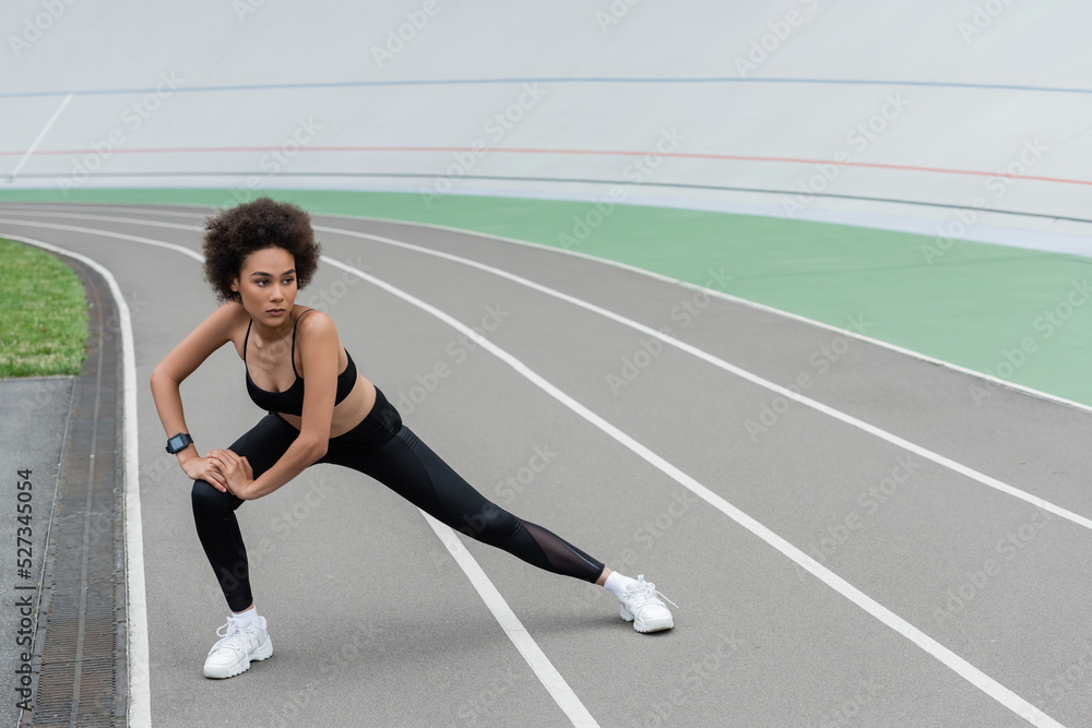 full length of african american woman in black sportswear doing side lunge exercise while stretching on stadium. full length of african american woman in black sportswear doing side lunge exercise while stretching on stadium.