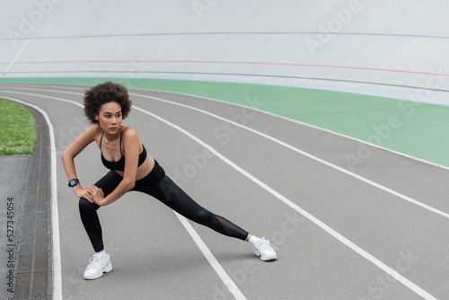 full length of african american woman in black sportswear doing side lunge exercise while stretching on stadium.