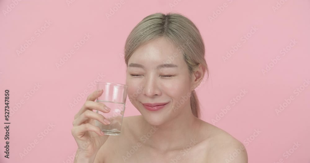 Studio portrait of a young, beautiful Asian woman with blond hair holding glass, then drinking water and smiling at the camera in pink background. Blogger with beauty, healthy or fashion concept. Studio portrait of a young, beautiful Asian woman with blond hair holding glass, then drinking water and smiling at the camera in pink background. Blogger with beauty, healthy or fashion concept.