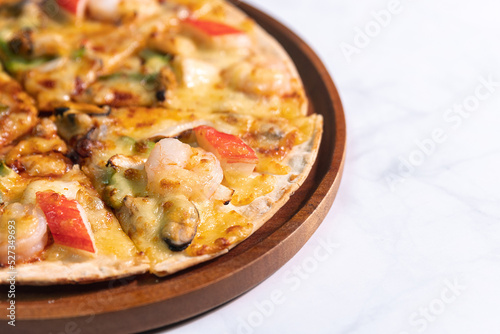 Close up of tasty hot baked seafood crispy pizza - mussels, shrimps and kani with chili pepper and melted mozzarella cheese on round pizza dough, on a wooden pizza pan over the marble table top.