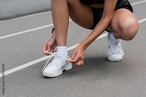 Cropped view of african american sportswoman tying shoelaces of white sneaker on stadium.