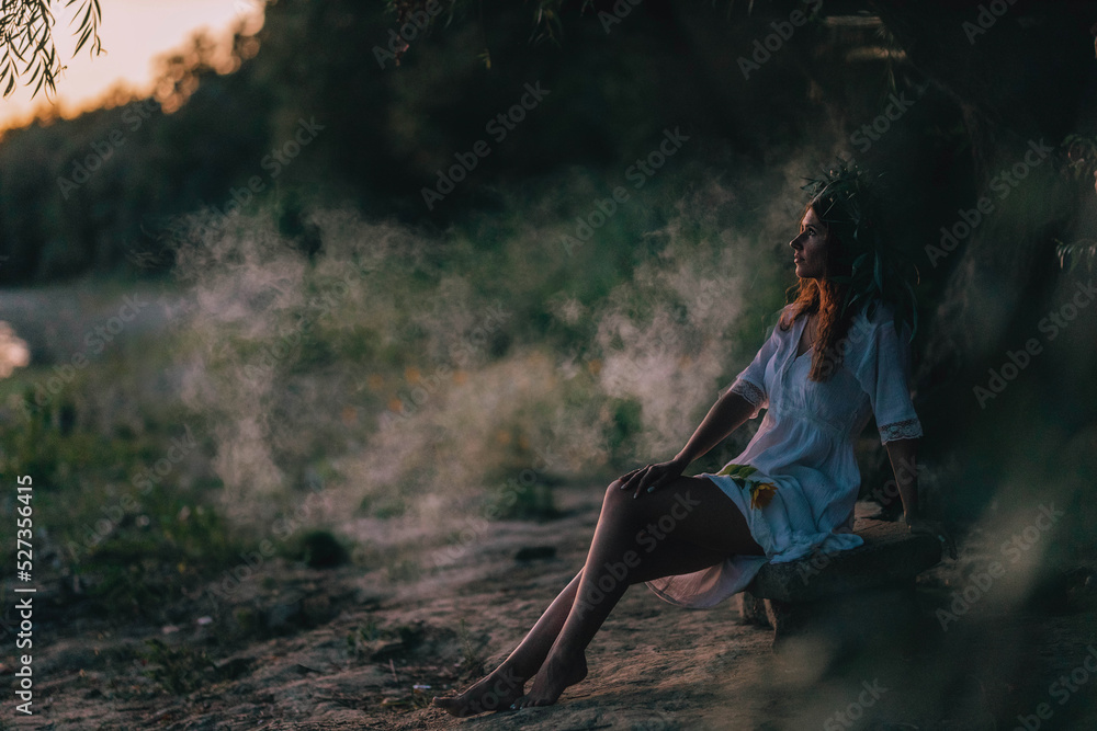 Girl in a white vintage dress with willow and flowers wreath sitting and dreaming near the river on stones . Harmony and rest with nature. Ivan Kupala tradition in Ukraine.  Fog is outside