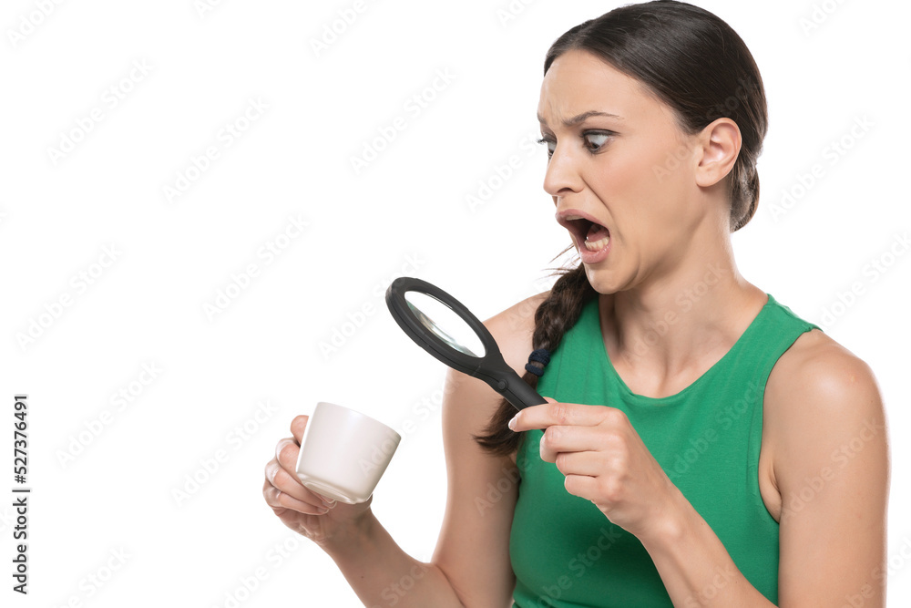 Shocked young woman looking with a magnifying glass in a cup of hot drink on a white background. Shocked young woman looking with a magnifying glass in a cup of hot drink on a white background.