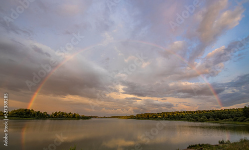 Picturesque rural landscape. Rainbow against a dramatic sky. Evening by the river.