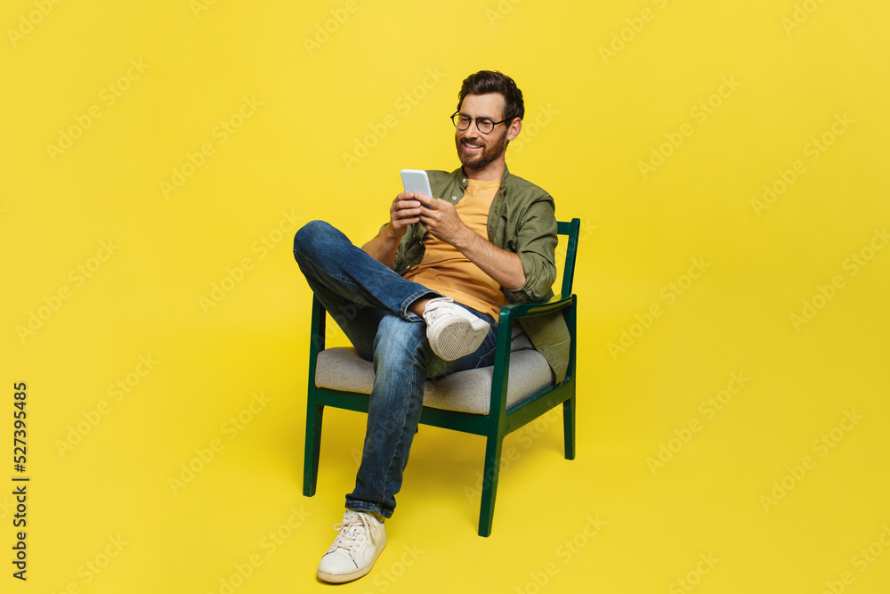Excited middle aged man using cellphone, networking in social media or texting, sitting in chair over yellow background