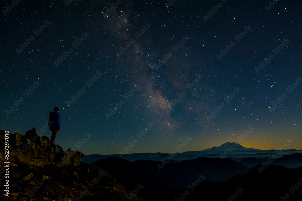 Athletic adventurous male hiker standing on top of a mountain looking out at the Milky Way and Mount Rainier. 