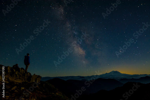 Athletic adventurous male hiker standing on top of a mountain looking out at the Milky Way and Mount Rainier. 