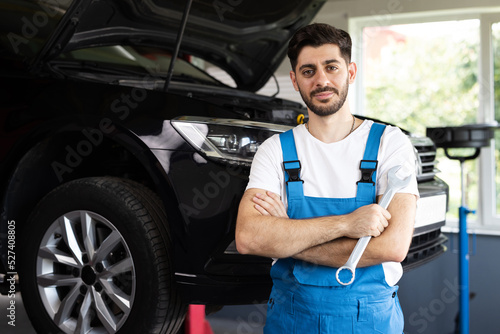 Confident handsome bearded mechanic in blue uniform standing with his arms crossed while workmate working on background. Car mechanic working at automotive service center © uflypro