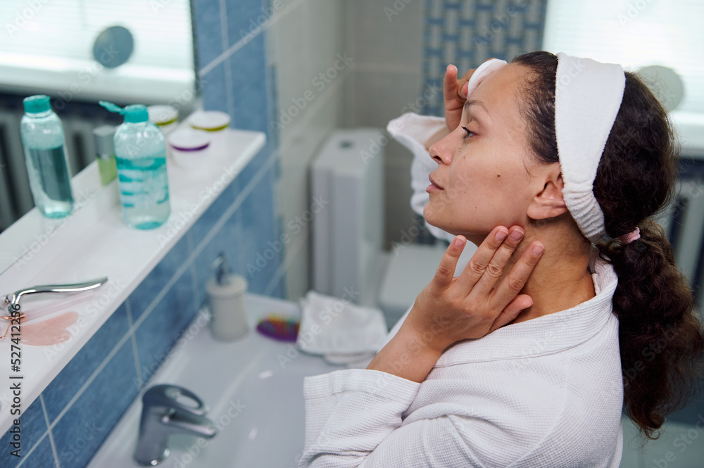 Close-up. Charming curly haired brunette middle-aged woman, applying cleansing lotion, to make her skin clear clean in front of a bathroom mirror, removes cosmetics from her eyes using a cotton pad Close-up. Charming curly haired brunette middle-aged woman, applying cleansing lotion, to make her skin clear clean in front of a bathroom mirror, removes cosmetics from her eyes using a cotton pad
