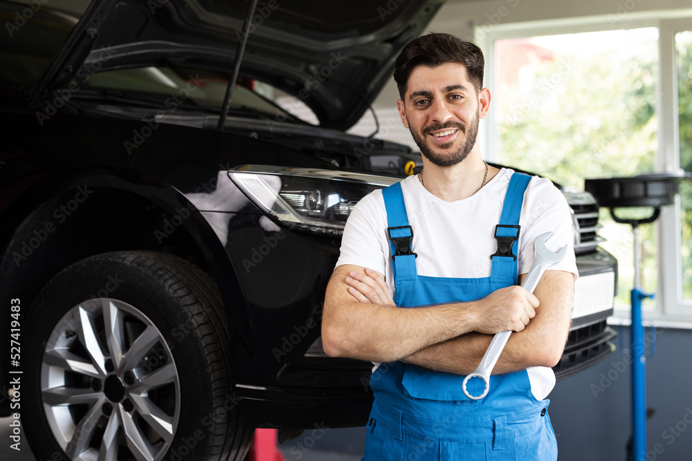 Young caucasian bearded man in blue coveralls holds spanner, smiling and looking into camera. Male car mechanic in spacious repair shop Young caucasian bearded man in blue coveralls holds spanner, smiling and looking into camera. Male car mechanic in spacious repair shop