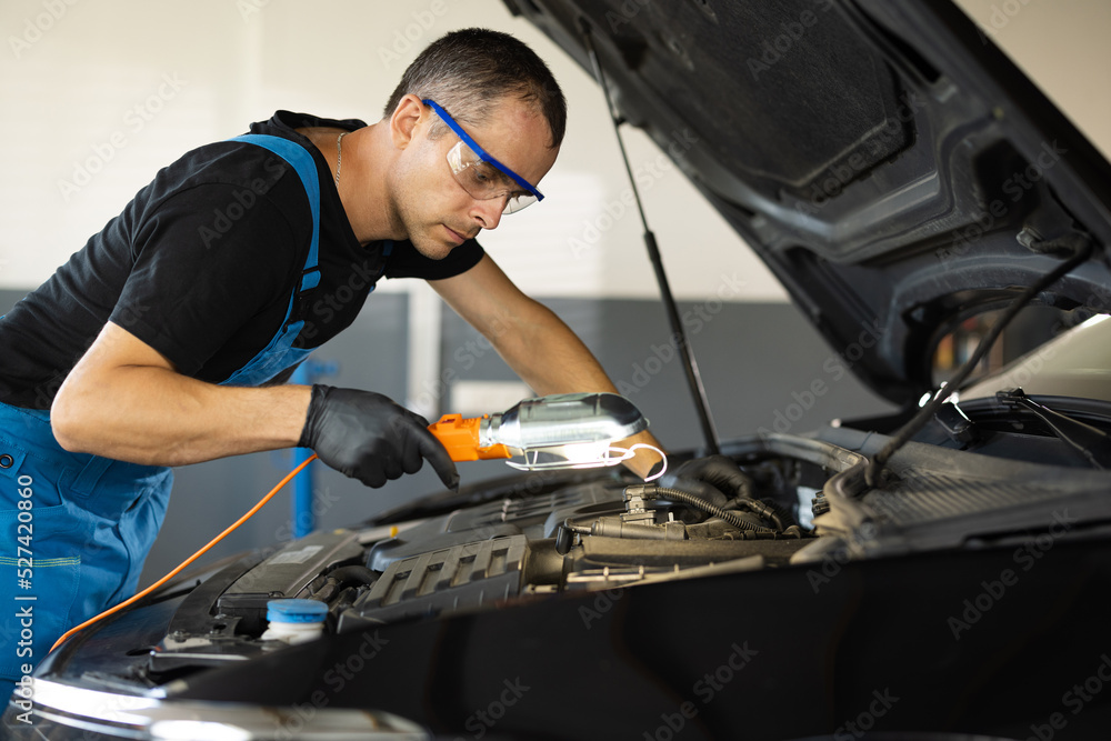 The mechanic in blue overalls and safety glasses inspects the car while working with led lamp. The work of a car mechanic in a modern workshop, car service. The mechanic in blue overalls and safety glasses inspects the car while working with led lamp. The work of a car mechanic in a modern workshop, car service.
