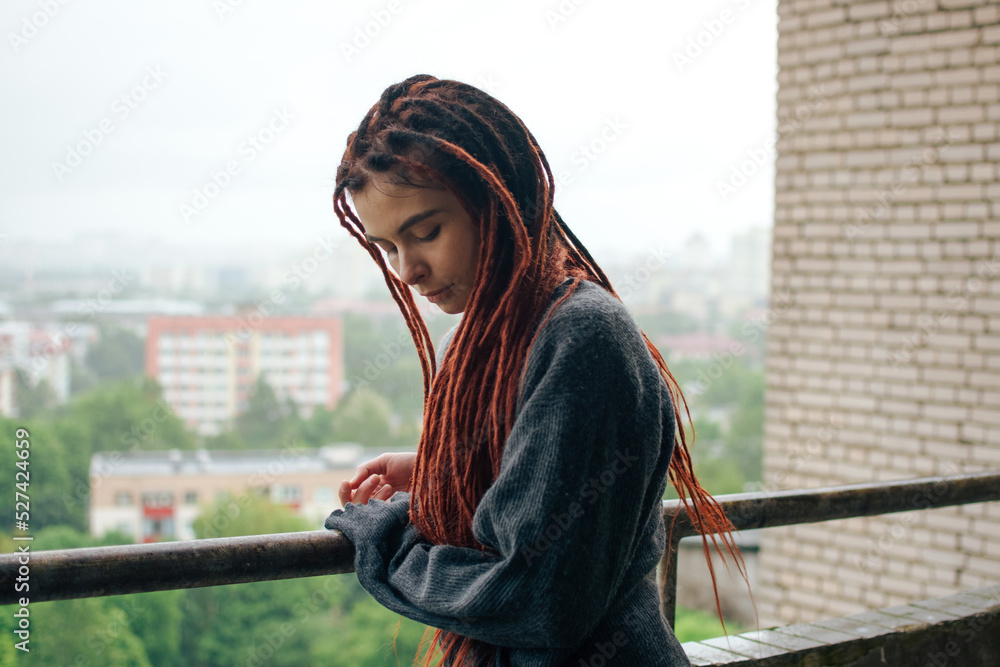 Young sad girl with red dreadlocks in a gray sweater looking at the city from the balcony after the rain. Sensual woman on the balcony. Autumn mood. 
