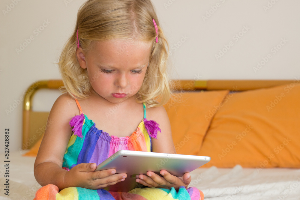 Little blond girl sitting on bed and holding tablet,toddler using internet with gadget,kid addicted to tablet and looking cartoons,3 year old child studiyng online with developmental games. Little blond girl sitting on bed and holding tablet,toddler using internet with gadget,kid addicted to tablet and looking cartoons,3 year old child studiyng online with developmental games.
