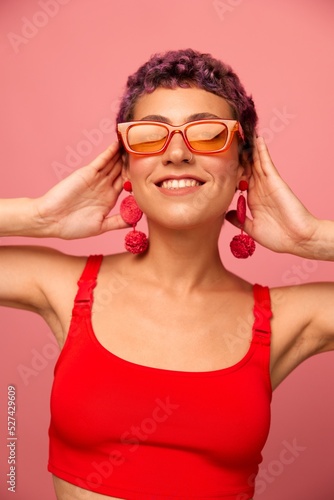 Young athletic woman with a short haircut and purple hair in a red top and pink yoga leggings in sunglasses with an athletic figure smiles and dances on a pink background
