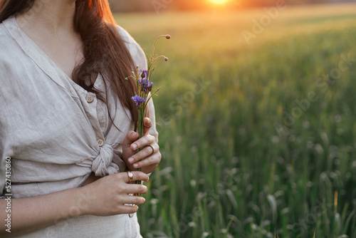 Woman holding wildflowers in wheat field in warm sunset light. Close up of young female in rustic dress with flowers in hands in evening summer countryside. Tranquil atmospheric moment
