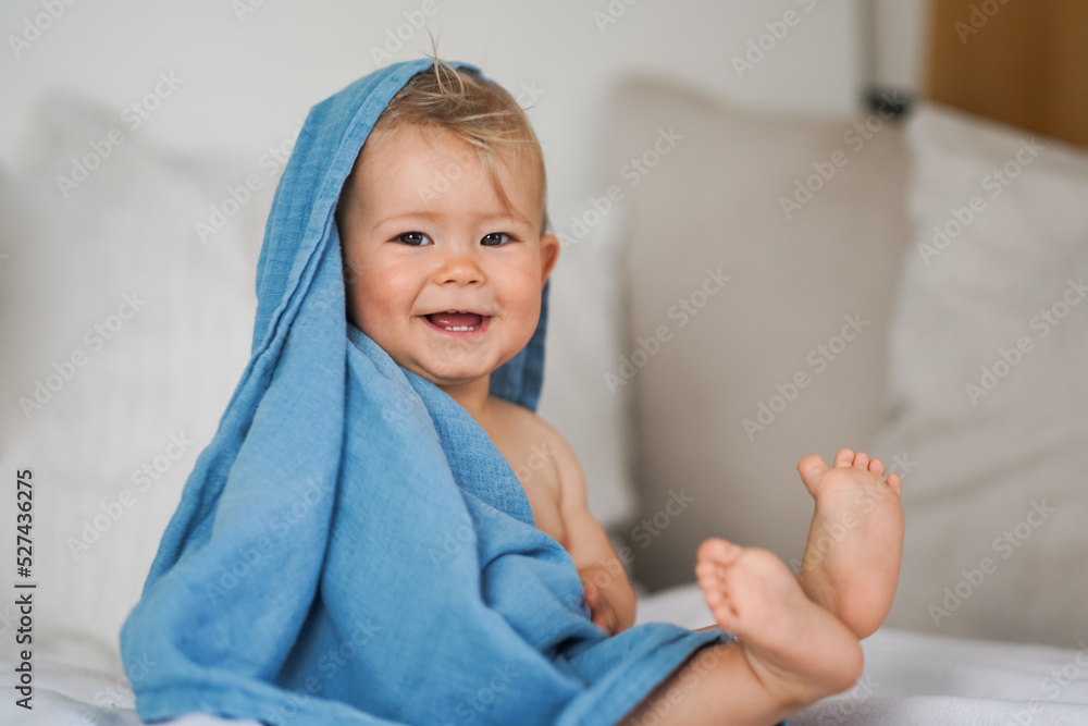 mega cute naked almost one year old blond baby boy sitting & laughing at home on a cozy bed after bathing and playing with a blue muslin fabric burp cloth while making nonsense jokes - obrazy, fototapety, plakaty 