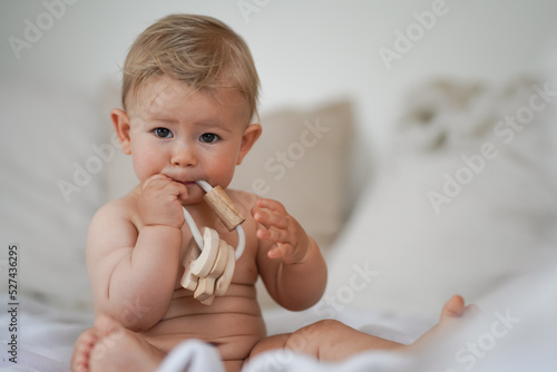 cute naked 10 month old blond baby boy sitting at home on a cozy bed after bathing and playing or biting a wooden eco teether toy because of teething