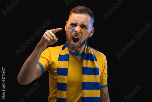 Portrait of excited young soccer supportred man in yellow-blue t-shirt and scarf and painted face, cheering for his favourite team, isolated over black background photo