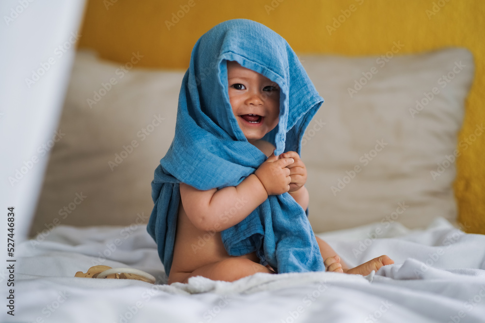 mega cute naked almost one year old blond baby boy sitting & laughing at home on a cozy bed after bathing and playing with a blue muslin fabric burp cloth while making nonsense jokes - obrazy, fototapety, plakaty 