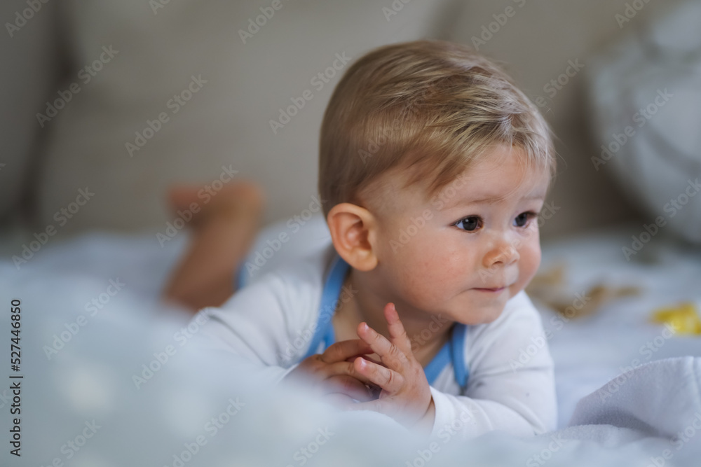 cute happy one year old happy blond babyboy in a white body or suit at home on a cozy bed after bathing and looking into the Camera cute happy one year old happy blond babyboy in a white body or suit at home on a cozy bed after bathing and looking into the Camera